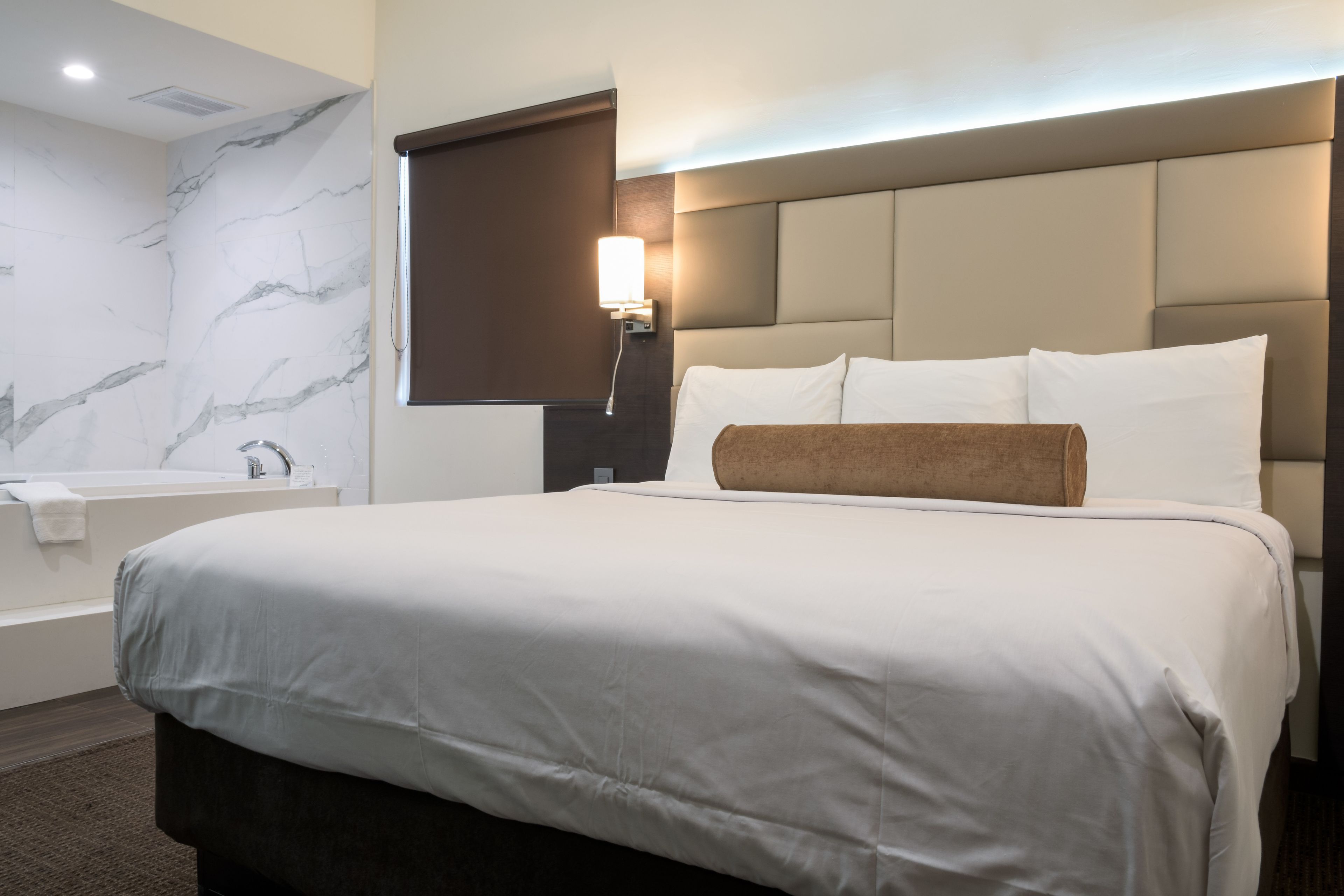 A modern hotel room with a large bed, a headboard, a wall-mounted light, and a bathroom with a bathtub and towel.