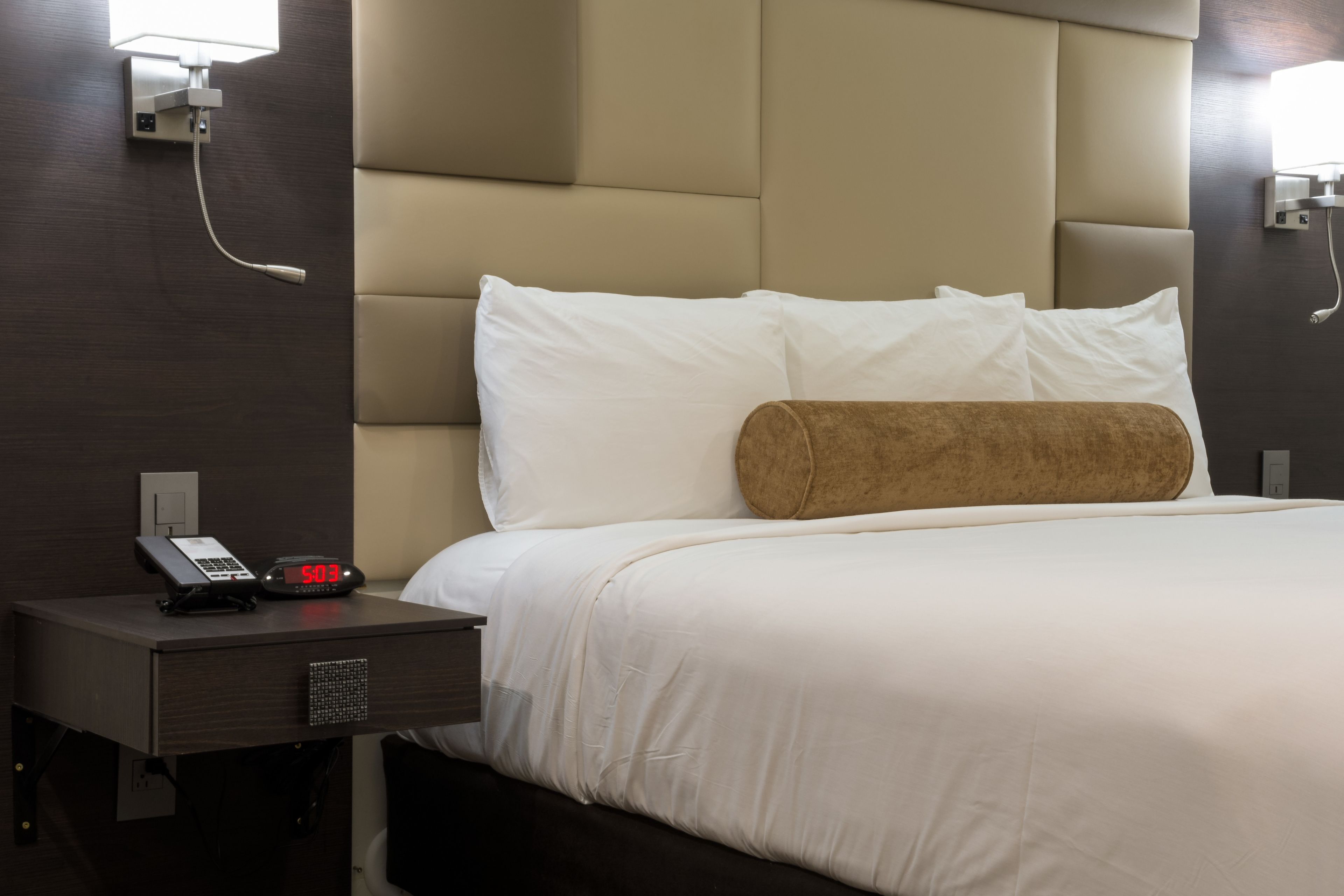 A hotel room with a bed, a nightstand with a phone and a digital clock, and a wall-mounted lamp.