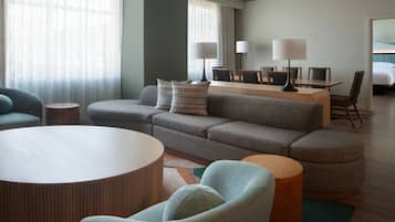 A modern hotel room with a grey sofa, a round coffee table, and a dining area with a wooden table and chairs.