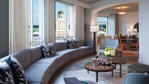 A modern hotel room with a curved gray sofa, a round coffee table with wine glasses, and a view of a clock tower through large windows. - Houston Marriott Sugar Land (Sugar Land)