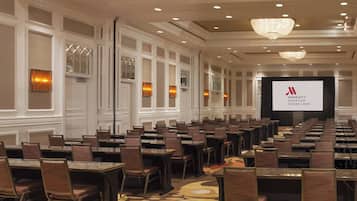 Una sala de conferencias con filas de mesas y sillas, una pantalla grande mostrando el logotipo de Marriott Houston Sugar Land y una lámpara de araña en el techo.