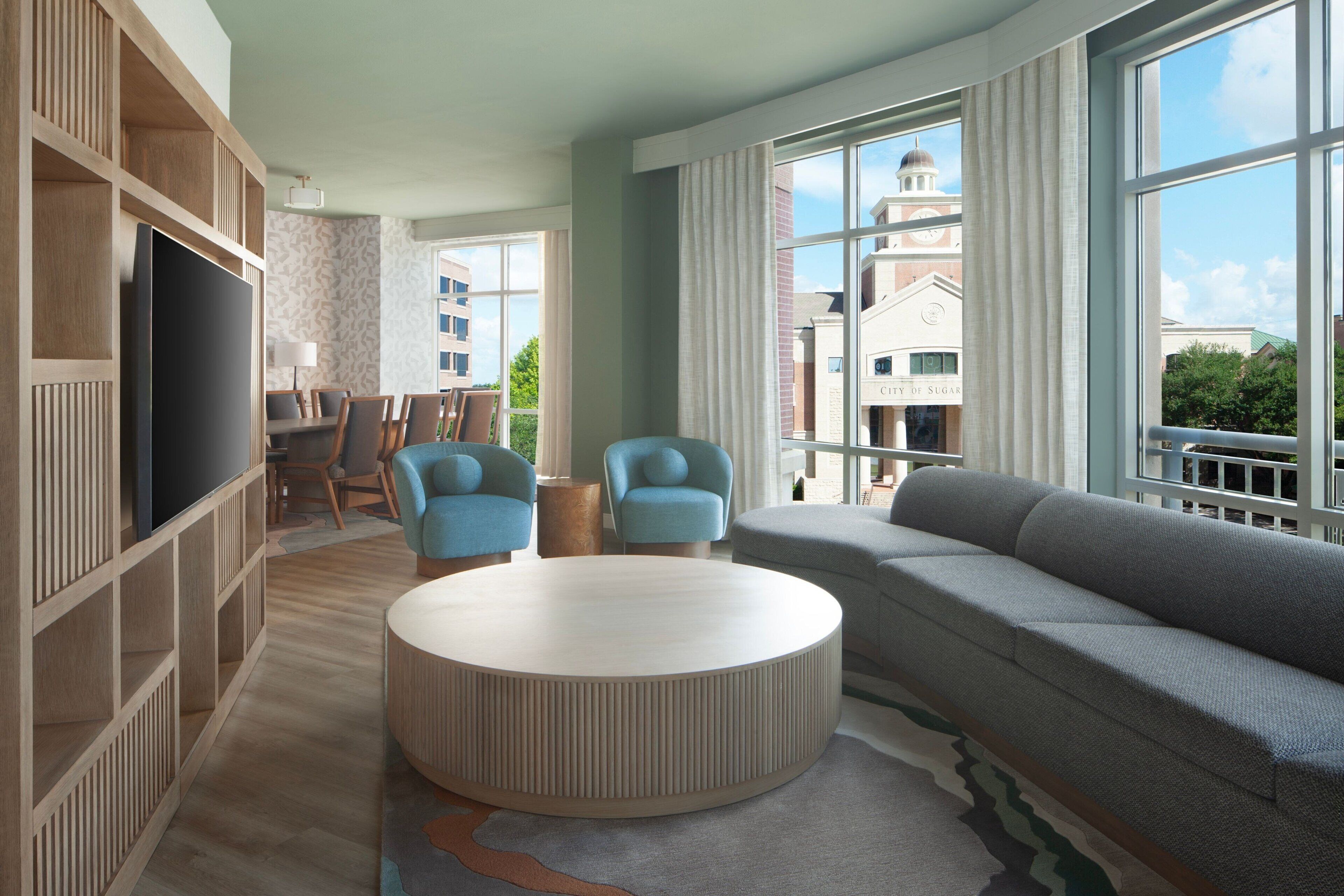 A modern living room with a large sofa, two armchairs, a round coffee table, and a TV on a wooden shelving unit.