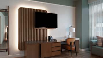 A modern hotel room with a wooden desk, a flat-screen TV, a chair, a sofa, and a window with sheer curtains.