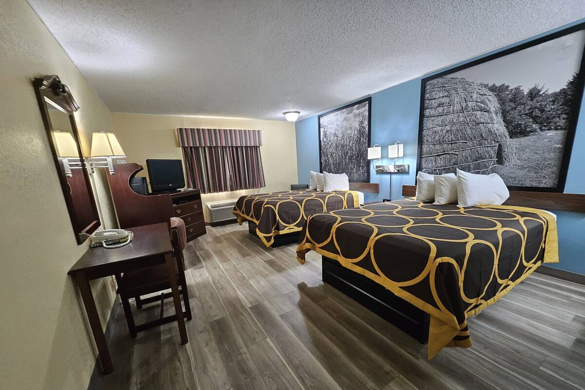 A hotel room with two beds, a desk, a chair, a TV, and a large window with a view of trees.