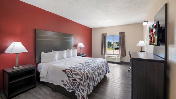 A hotel room with a bed, two bedside tables, a desk, a TV, and a window with curtains. - Red Roof Inn & Suites Austin East - Manor (Manor)