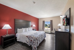A hotel room with a bed, two bedside tables, a desk, a TV, and a window with curtains. - Red Roof Inn & Suites Austin East - Manor (Manor)