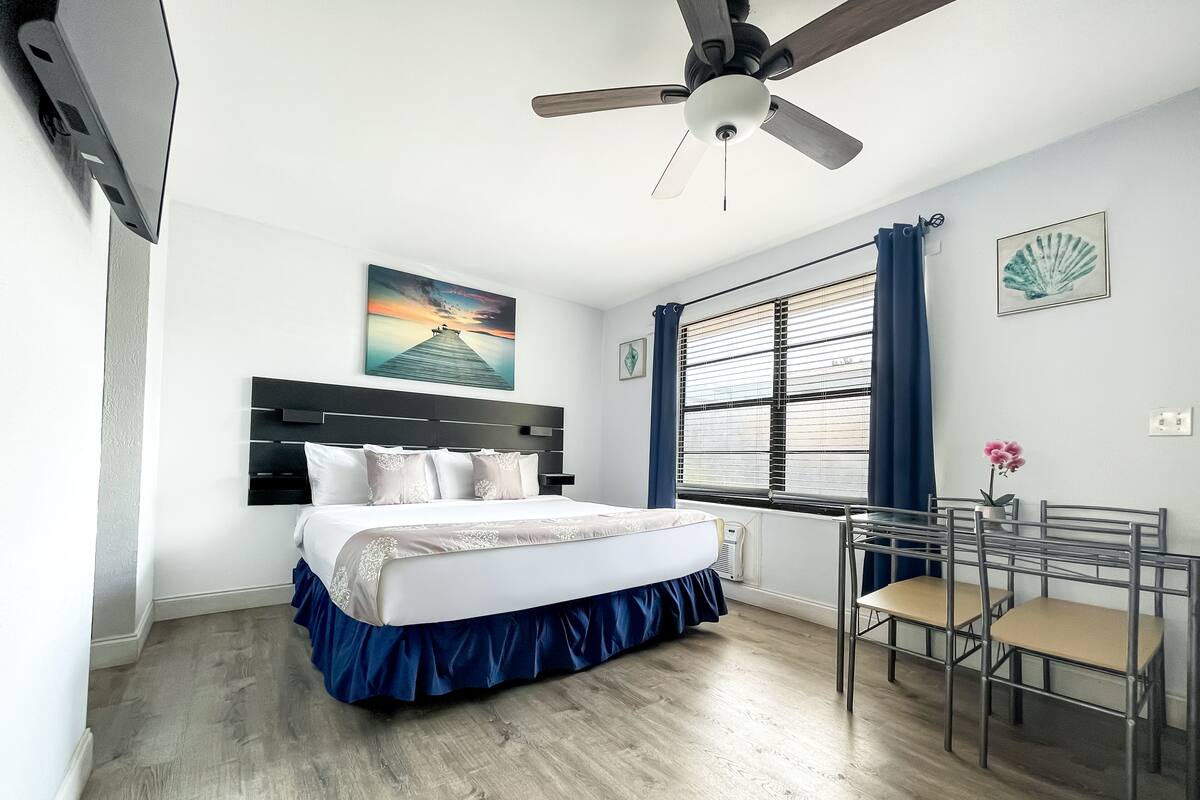 A bedroom with a bed, a ceiling fan, a window with blinds, a desk with chairs, and a painting on the wall.