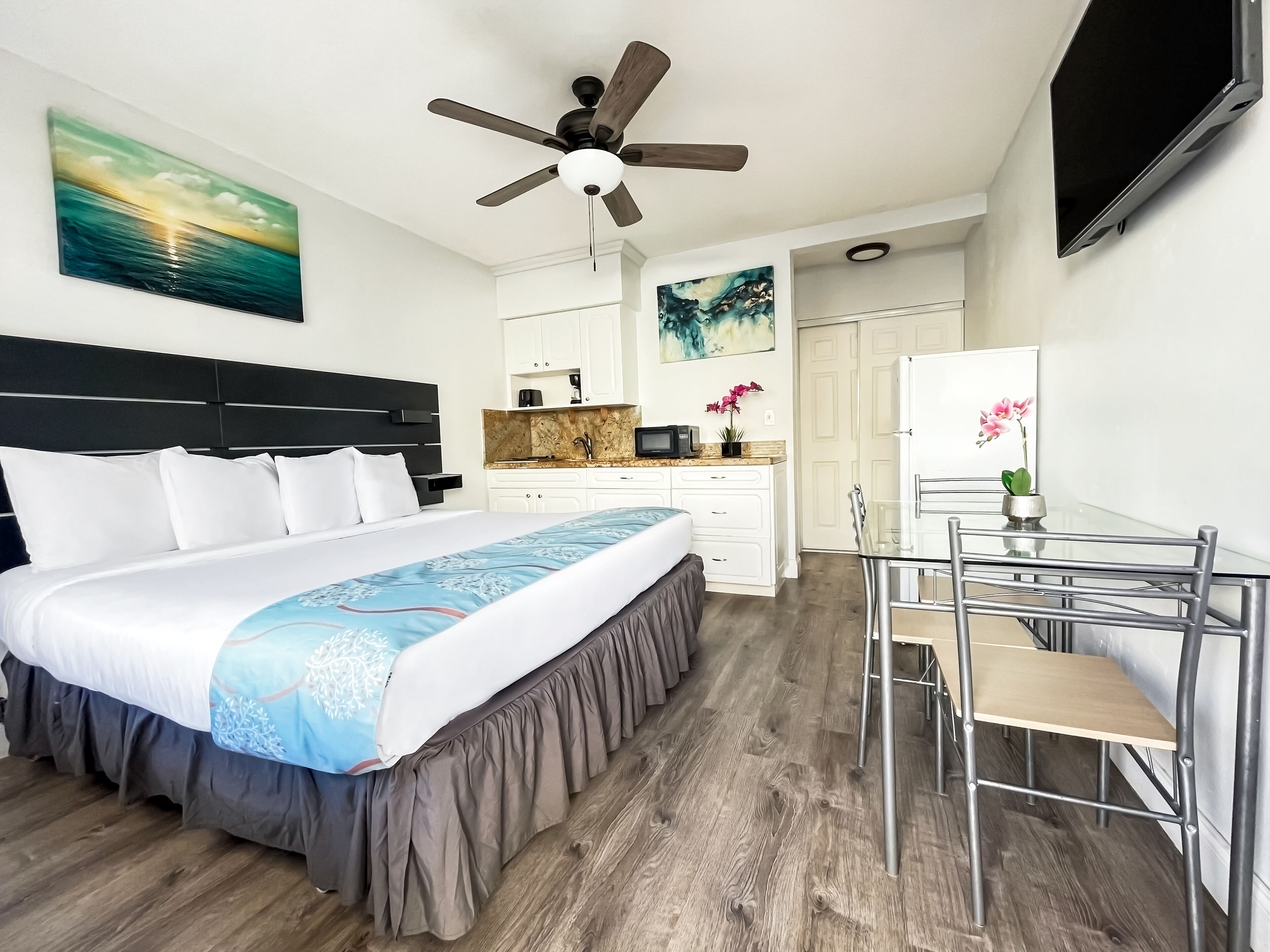 A hotel room with a bed, a desk, a chair, a ceiling fan, a TV, and artwork on the walls.