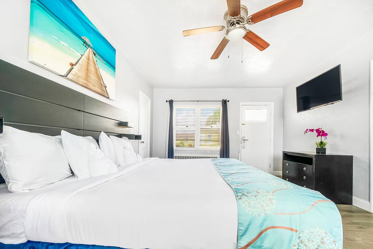 A bedroom with a bed, a ceiling fan, a TV, and a window with blinds.