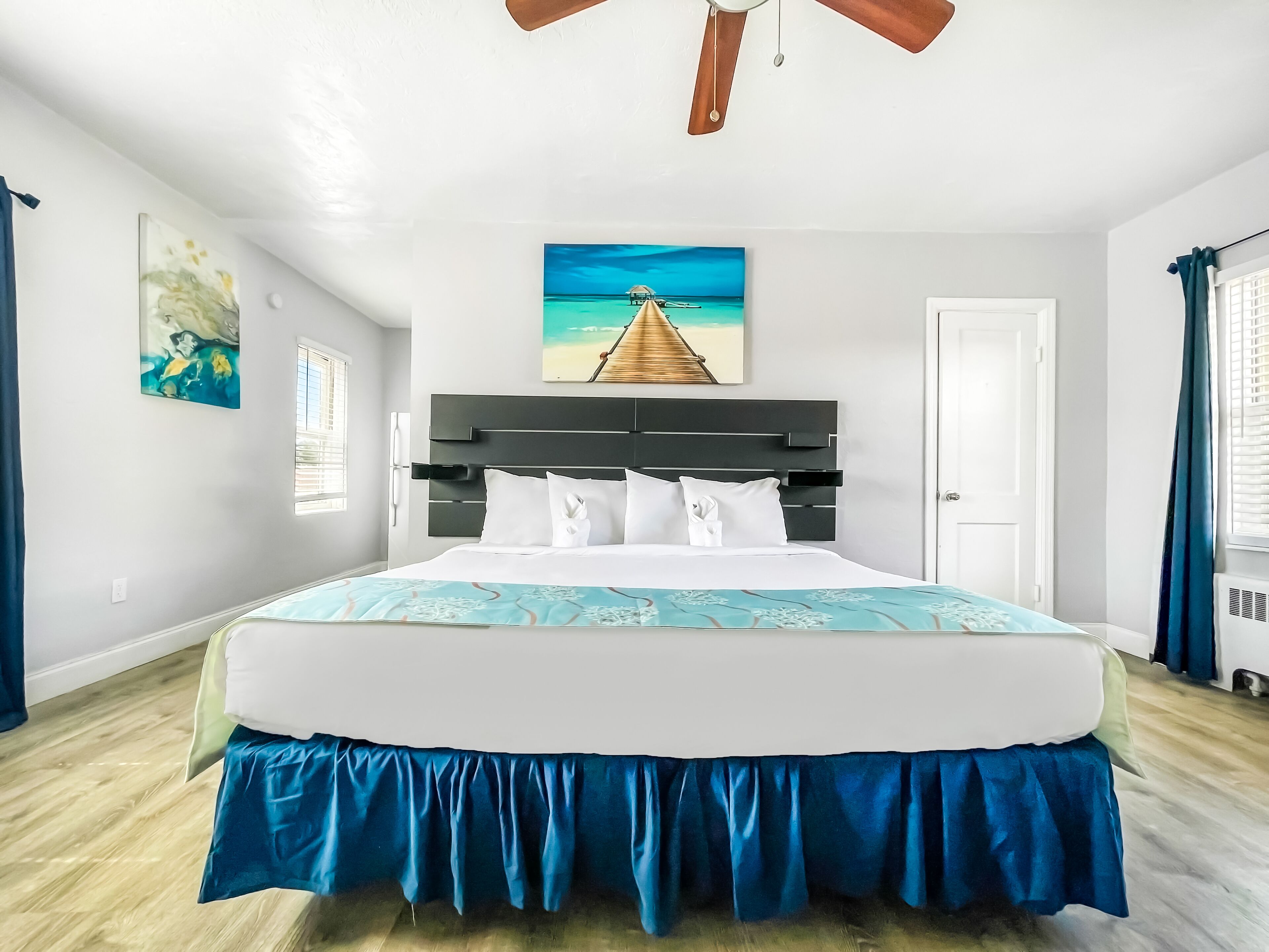 A hotel room with a large bed, a ceiling fan, a painting of a beach scene, and a window with curtains.