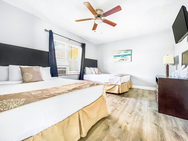 A hotel room with two beds, a ceiling fan, a painting, and a desk with a lamp.