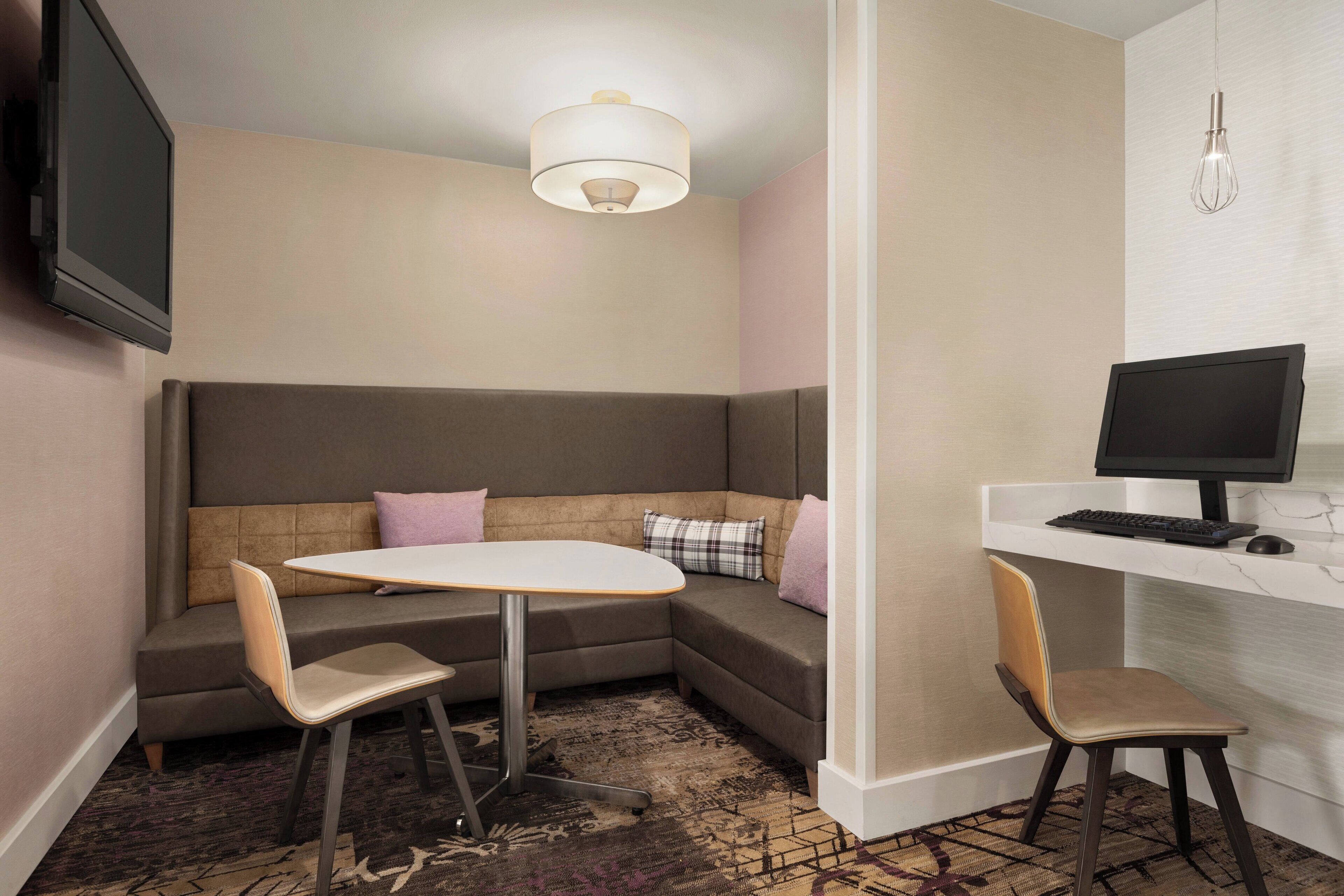 Una habitación de hotel moderna con un sofá modular, una mesita redonda pequeña, dos sillas, un escritorio con ordenador y un televisor de pantalla plana montado en la pared.