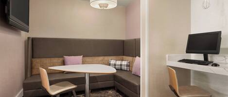 A modern hotel room with a sectional sofa, a small round table, two chairs, a desk with a computer, and a flat-screen TV mounted on the wall.