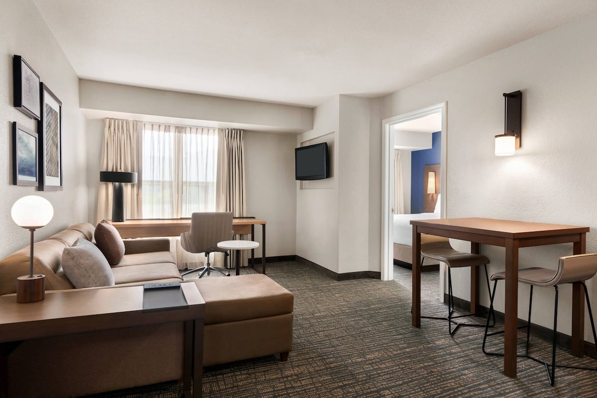 A hotel room with a sofa, a small table, a desk, a chair, a lamp, and a TV on the wall.