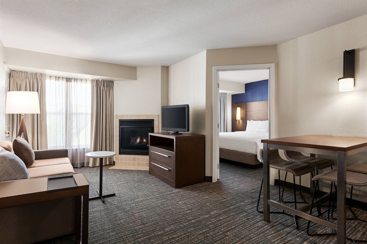 A hotel room with a sofa, a small table, a TV on a cabinet, a fireplace, and a separate bedroom area with a bed and a bathroom visible through an open door.