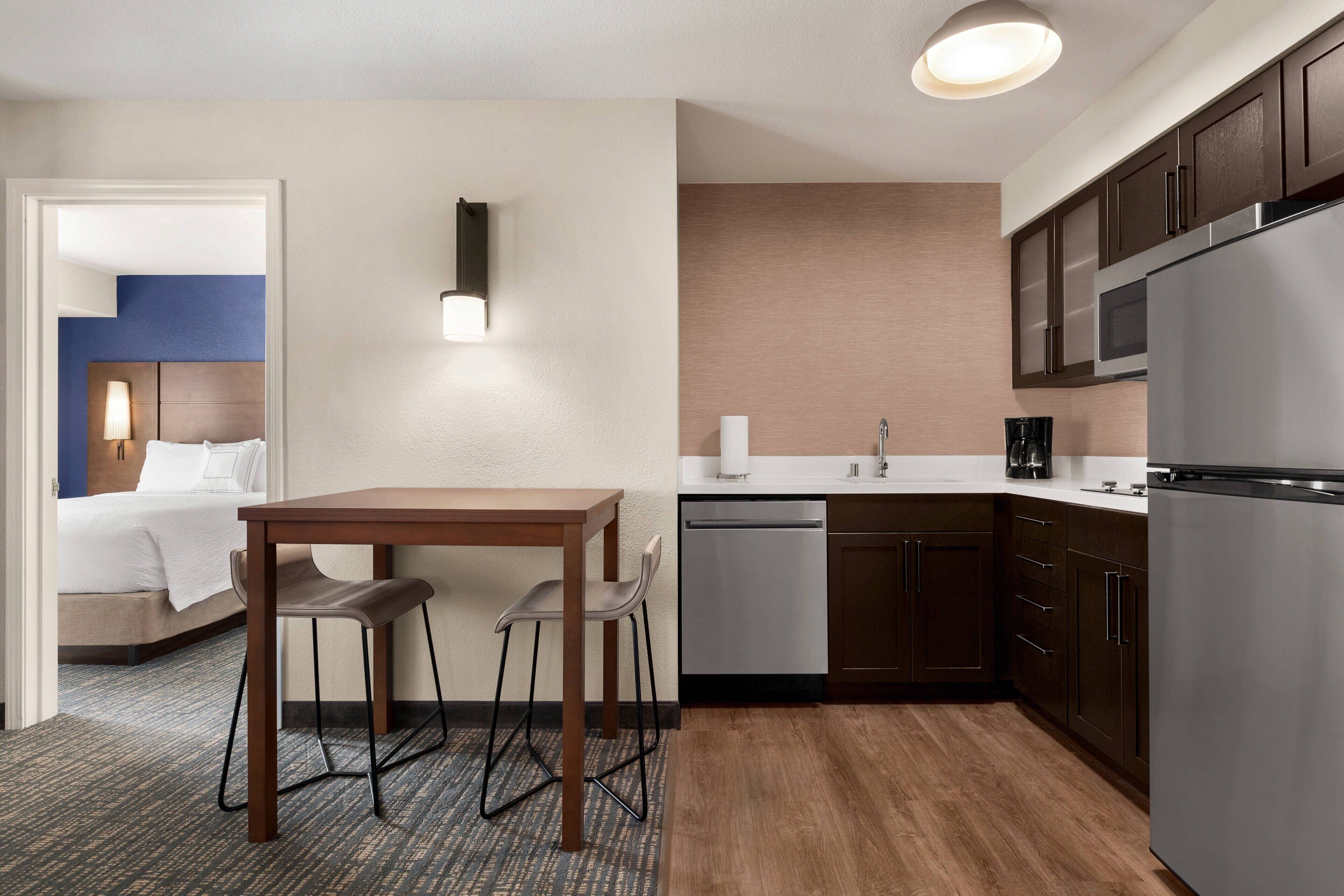 A compact hotel room with a kitchenette, a bed with white linens, a small dining table with two chairs, and a wall-mounted light fixture.