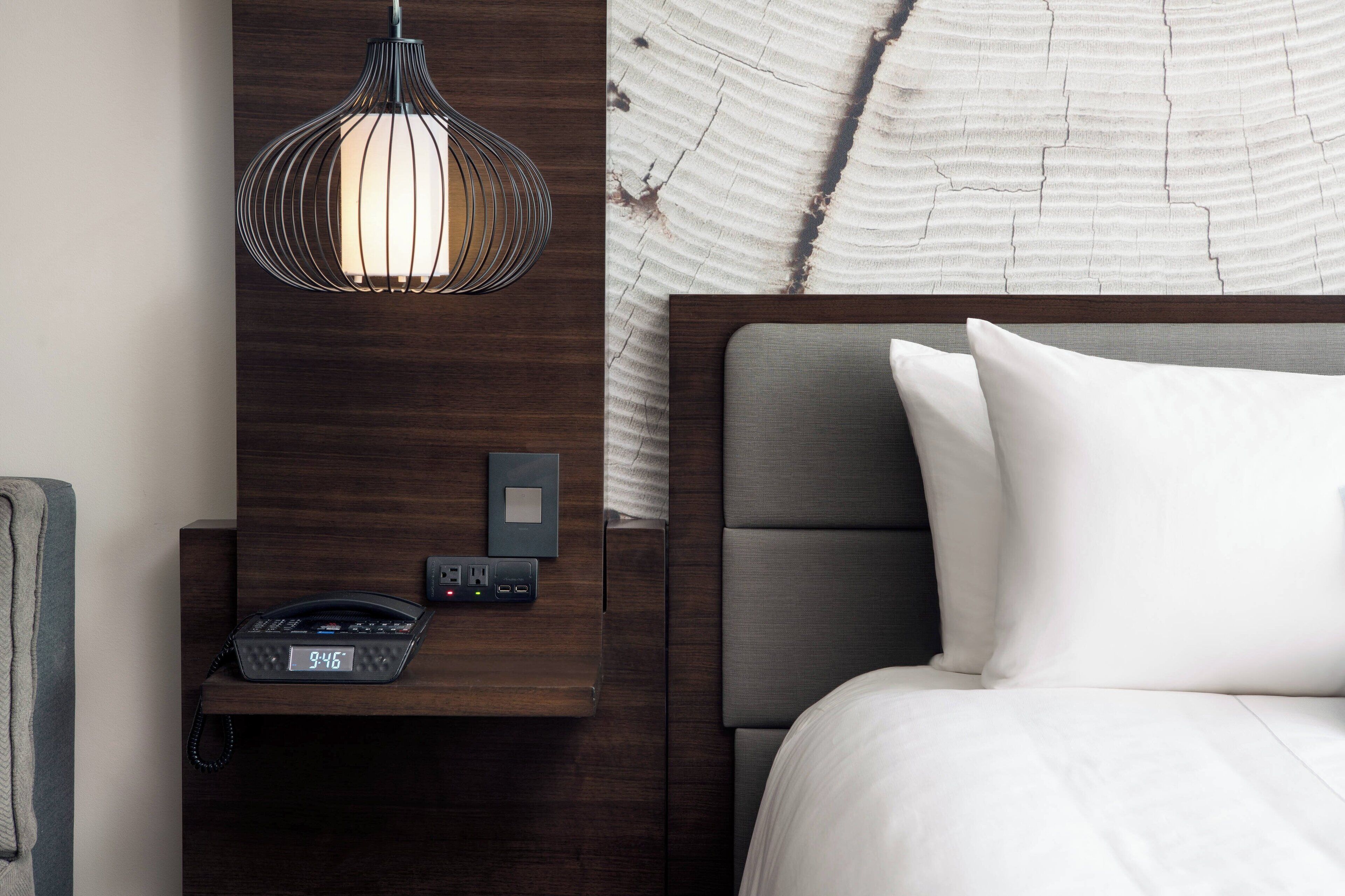 A hotel room with a wooden nightstand, a modern lamp, a digital clock, and a bed with white bedding.