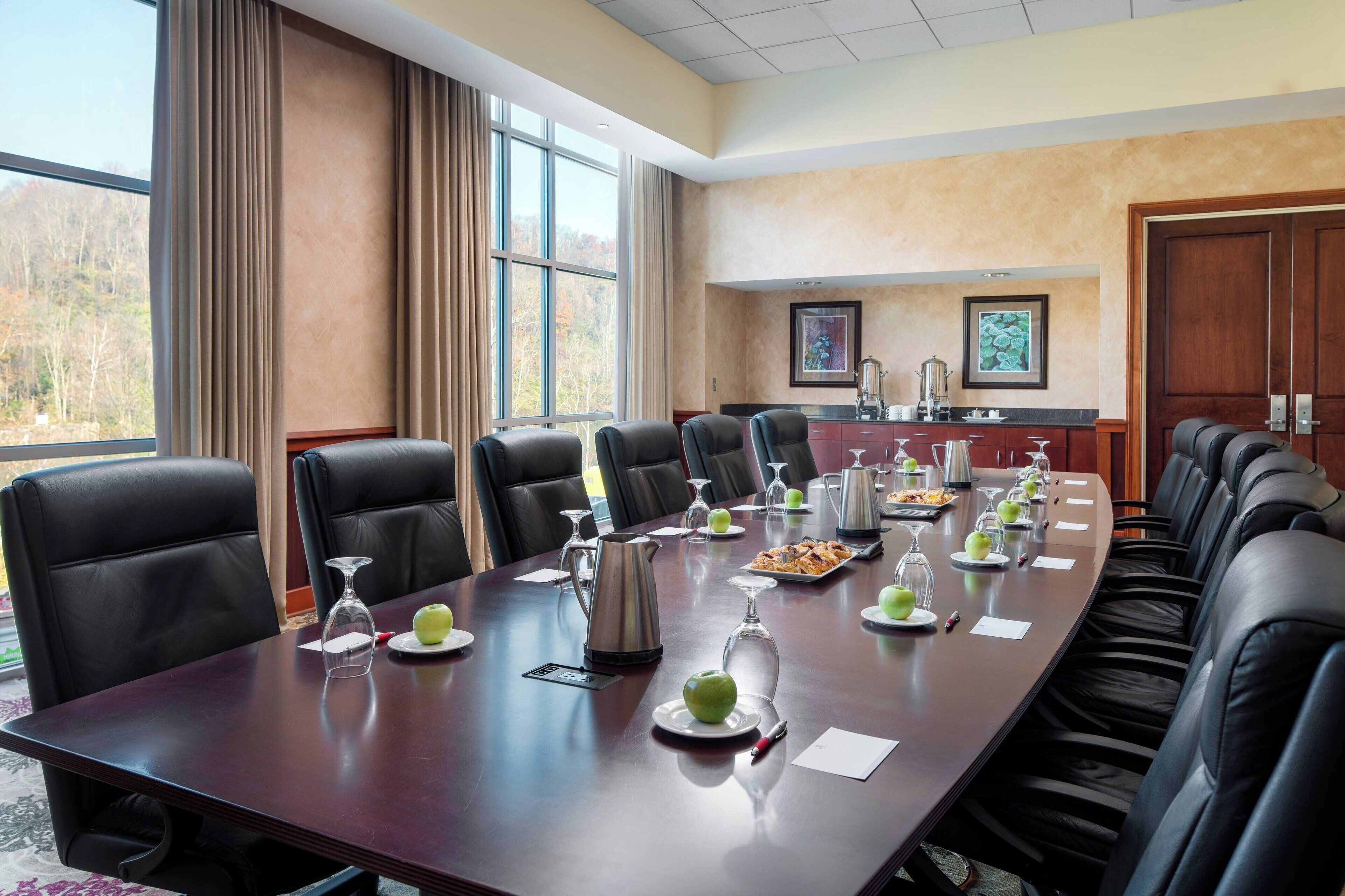 A conference room with a long table, black leather chairs, and a view of trees through large windows.