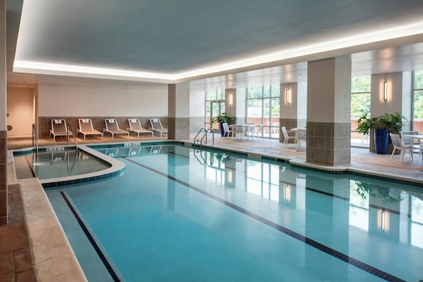 An indoor swimming pool with lounge chairs and tables, surrounded by large windows and potted plants.