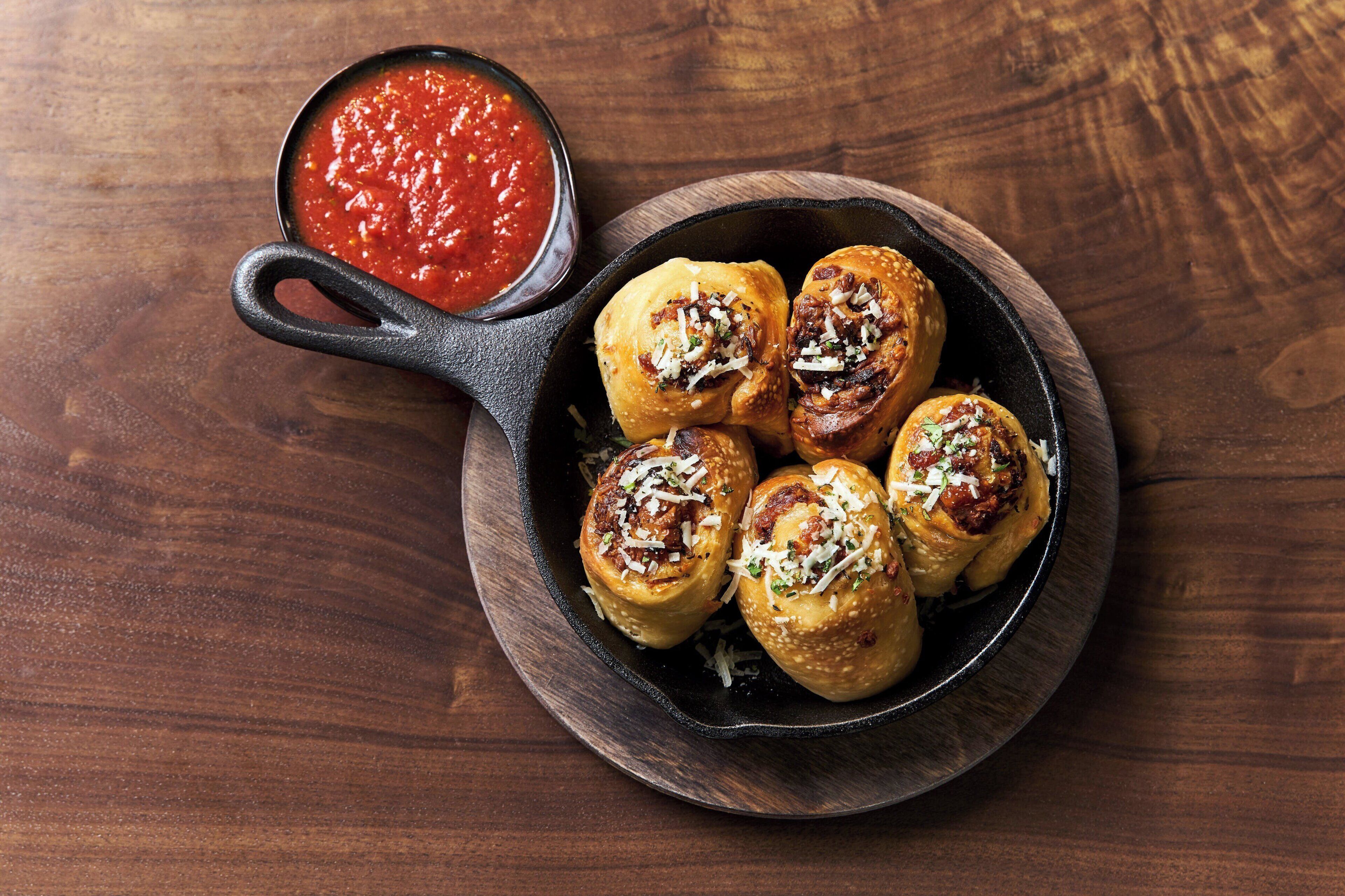 A skillet with six stuffed buns and a small bowl of red sauce on a wooden table.
