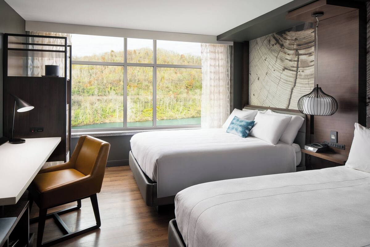 Ein modernes Hotelzimmer mit zwei Betten, einem Schreibtisch, einem Stuhl und einem großen Fenster mit Blick auf einen Wald.