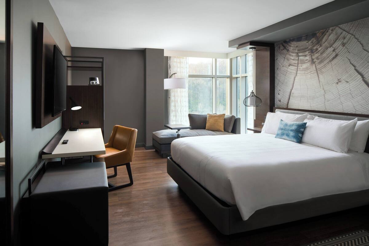 Ein modernes Hotelzimmer mit einem großen Bett, einem Schreibtisch und einem Stuhl. Ein Fenster mit Vorhängen und eine Karte an der Wand.