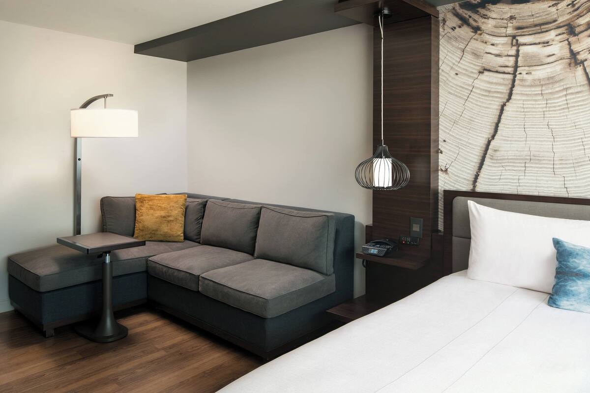 Ein modernes Hotelzimmer mit Ecksofa, kleinen Beistelltisch, Lampe, Pendelleuchte und einem Bett mit Kissen.