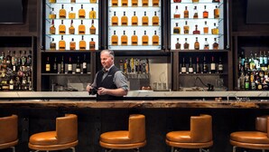 A bar with multiple shelves of liquor bottles, a bartender, and leather bar stools. - Morgantown Marriott at Waterfront Place (Morgantown)
