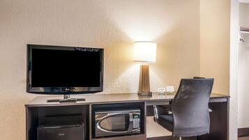 A hotel room with a flat-screen TV, a microwave, a desk with a lamp, and a chair.