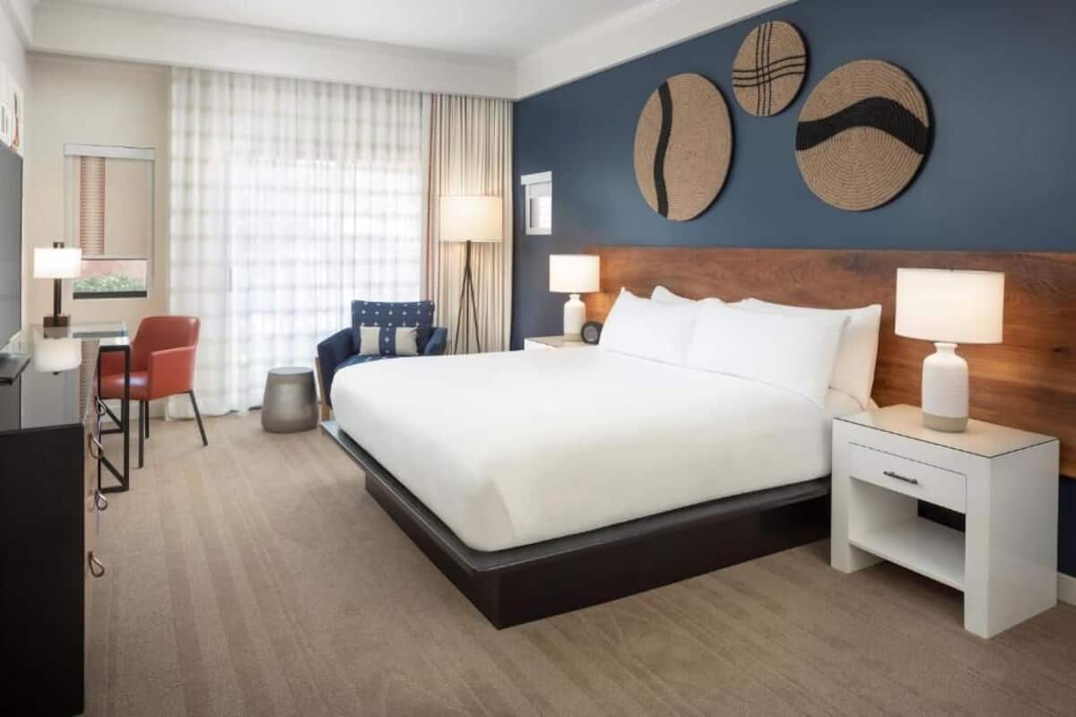 A hotel room with a large bed, two bedside tables with lamps, a desk with a chair, and a large window with curtains.
