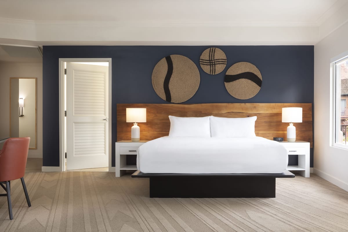 A hotel room with a large bed, two bedside tables with lamps, a wooden headboard, and a wall with three woven wall art pieces.