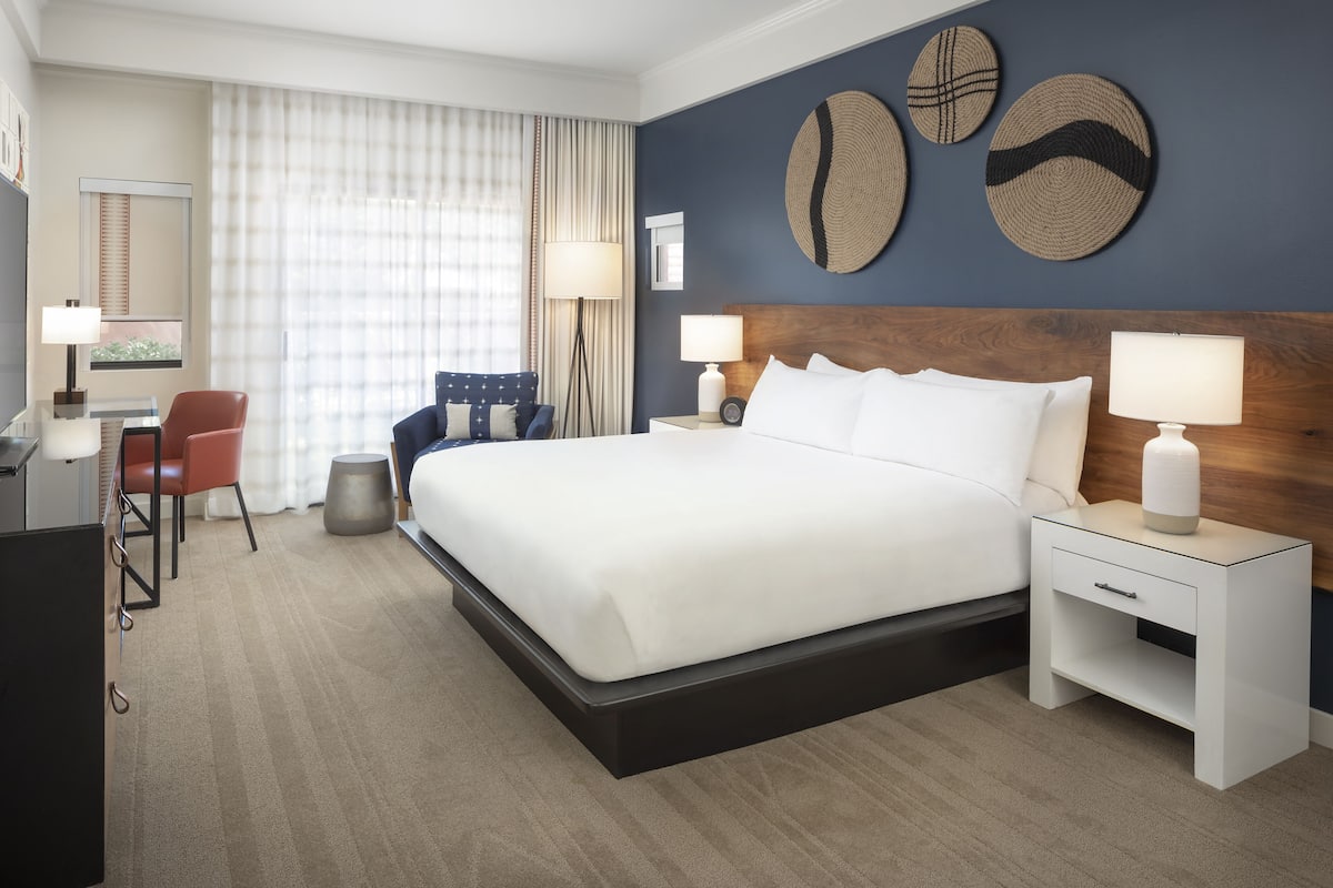 A hotel room with a large bed, two bedside tables with lamps, a desk with a chair, and a large window with curtains.