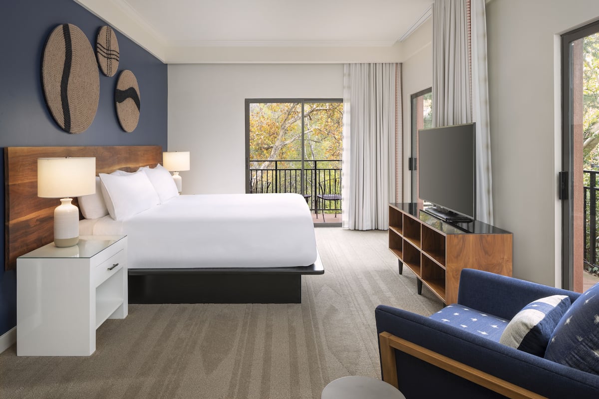 A hotel room with a large bed, a TV, a sofa, and a balcony with a view of trees.