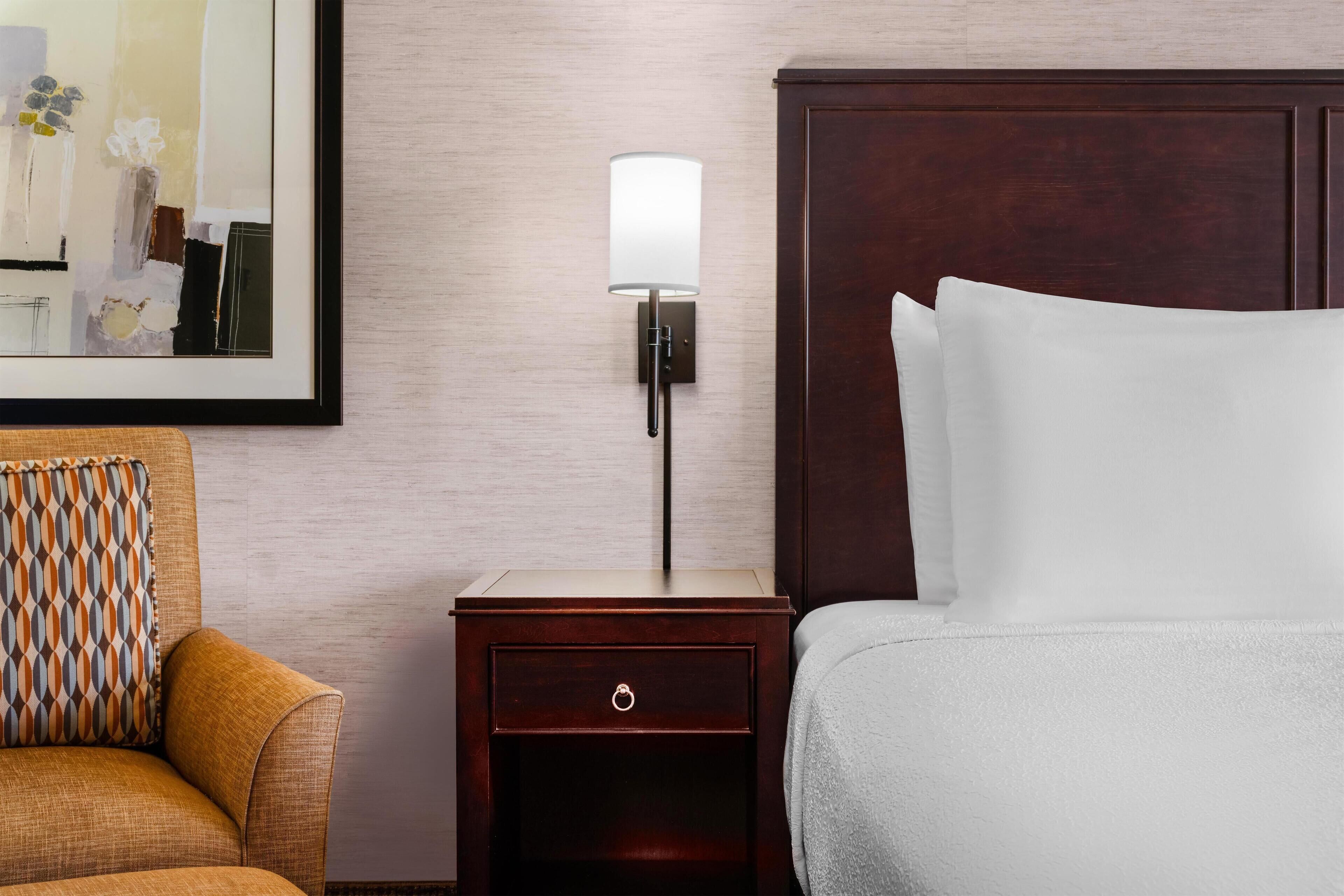 Camera d'albergo con un letto, un comodino con una lampada, una sedia e un quadro appeso al muro.