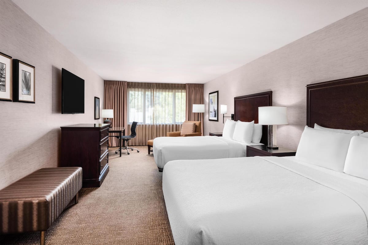 A hotel room with two beds, a desk, a chair, a bench, and a TV.