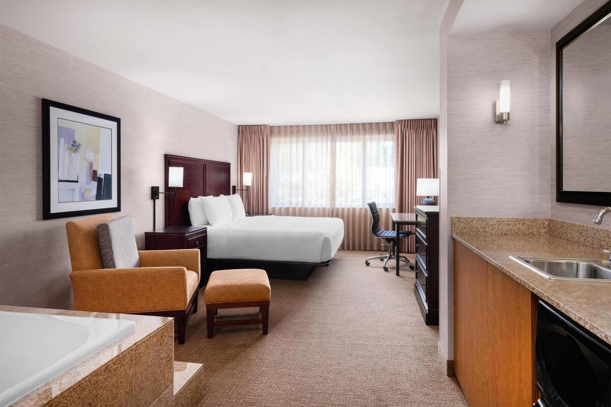 A hotel room with a bed, a desk, a chair, a small table, a sink, and a mirror.