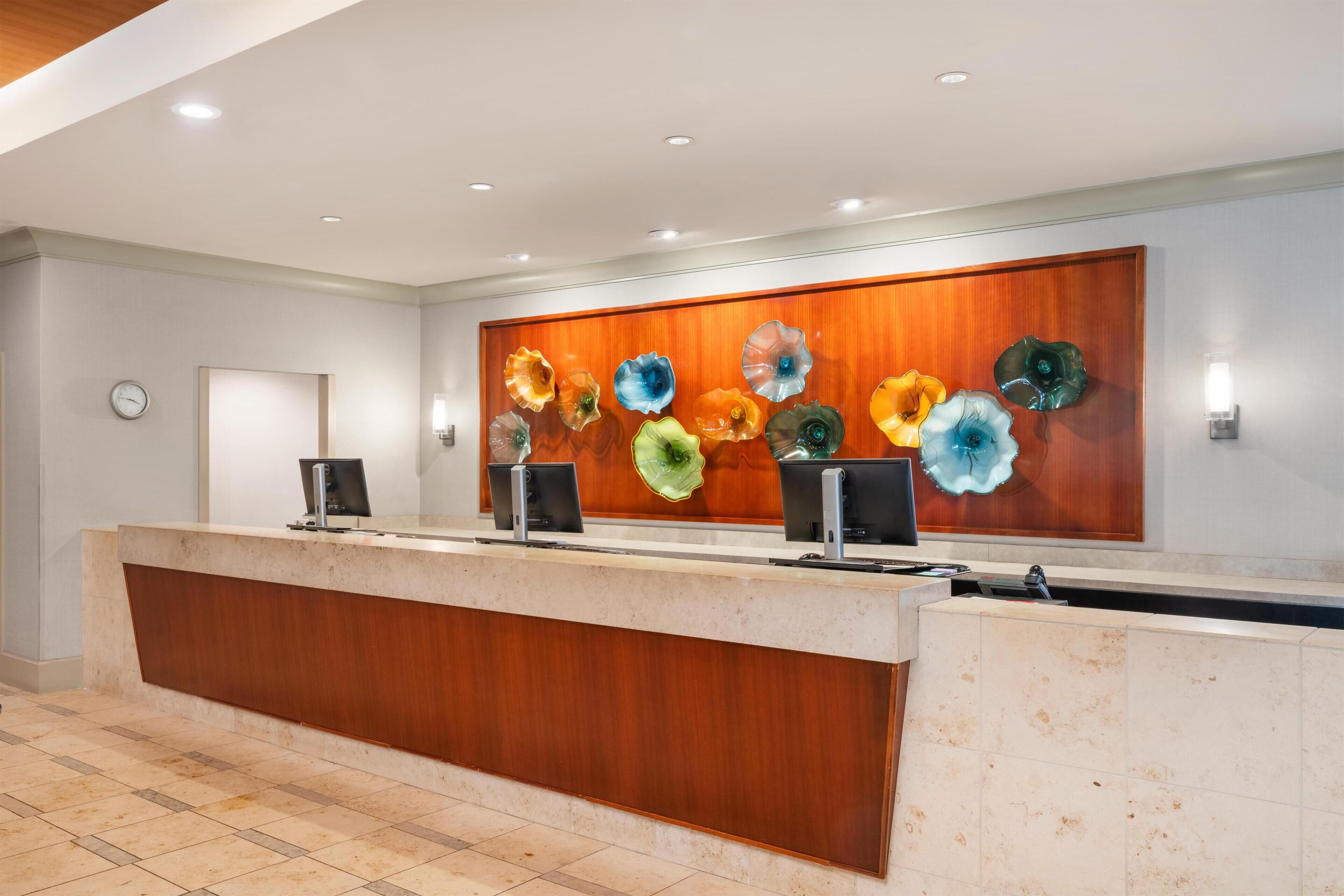 Zona de recepción de un hotel con un mostrador de madera, un reloj en la pared y una obra de arte de vidrio decorativa.