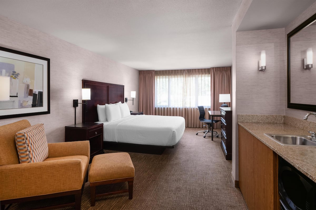 A hotel room with a bed, a desk, a chair, a sofa, and a kitchen area with a sink.