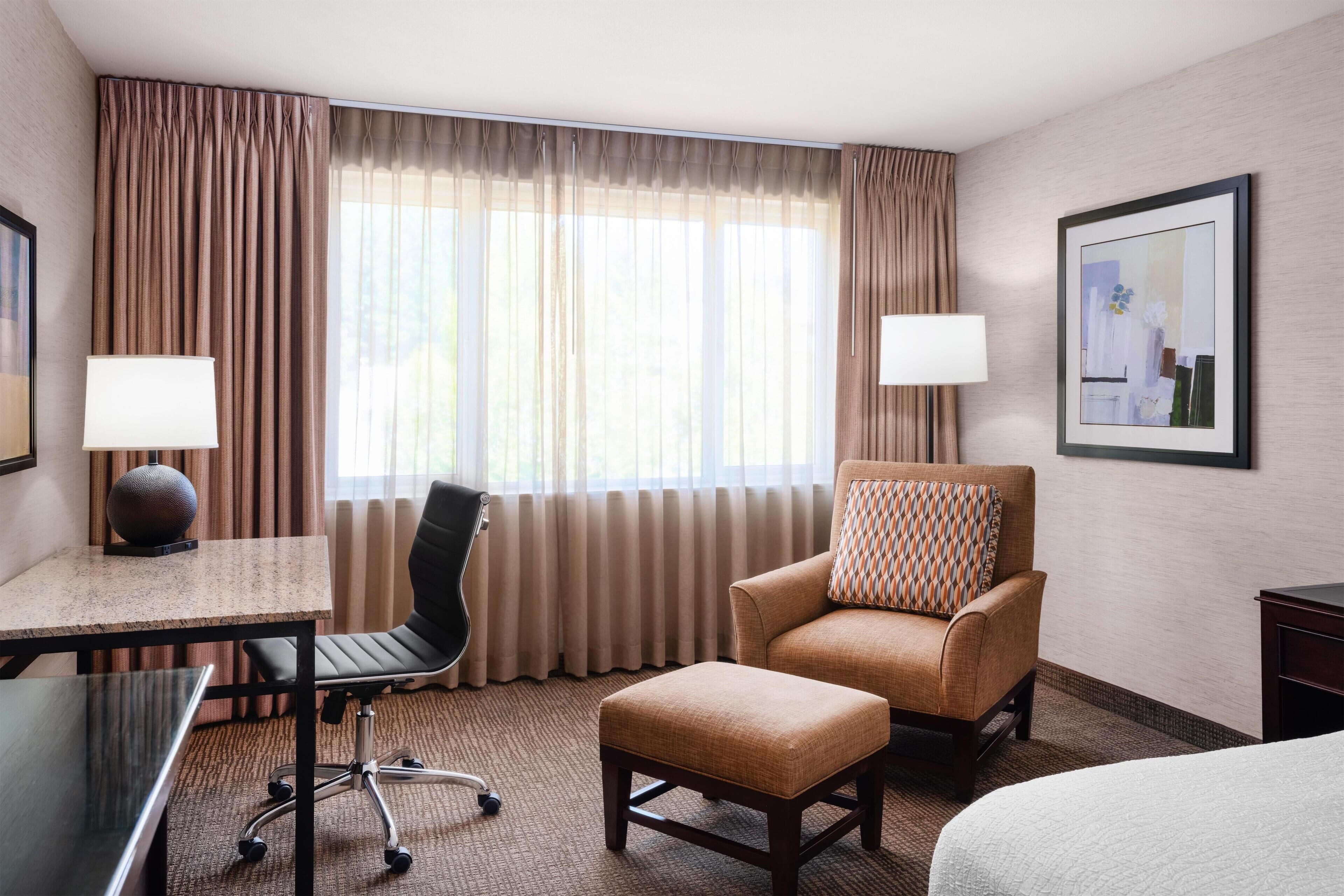 Een hotelkamer met een bed, een bureau met lamp, een stoel, een poef en een groot raam met gordijnen.