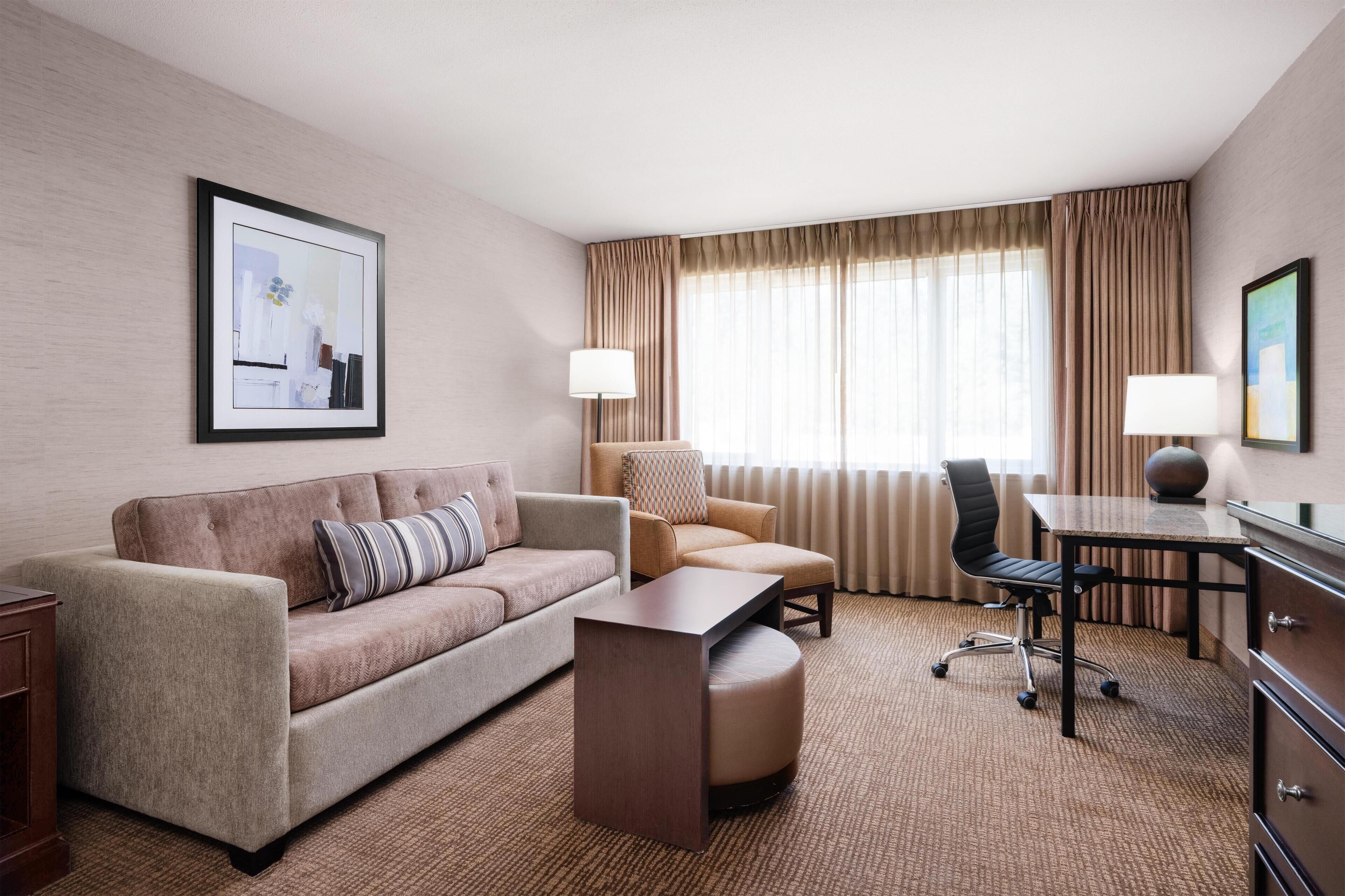A hotel room with a sofa, armchair, ottoman, desk, and lamps.
