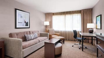 A hotel room with a sofa, armchair, ottoman, desk, and lamps.