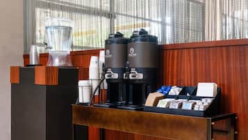 A coffee station with two coffee makers and a water dispenser, a stack of disposable cups, and a display of coffee pods.