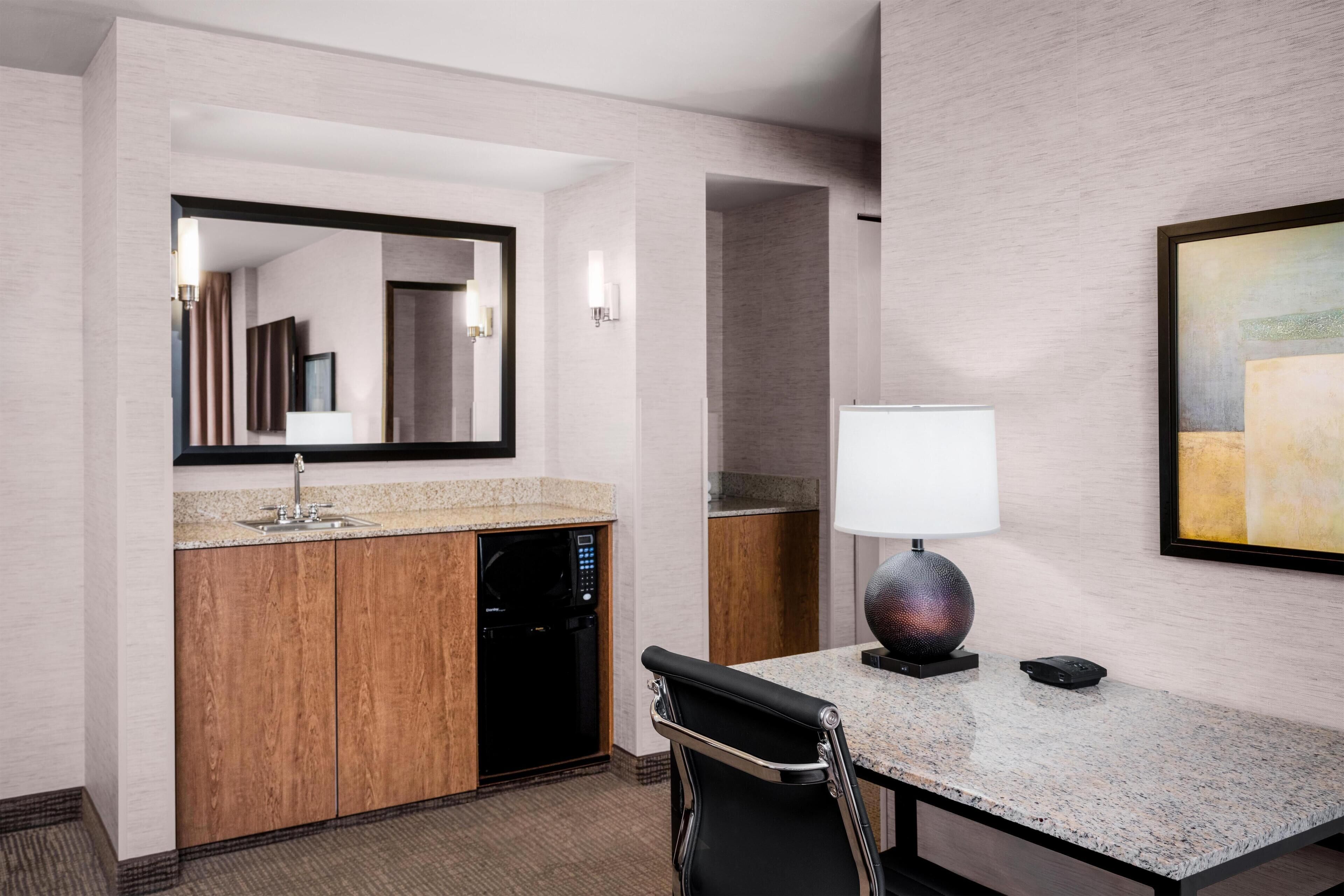 A modern hotel room with a kitchenette, a large mirror, a dining area with a table and chair, and a painting on the wall.