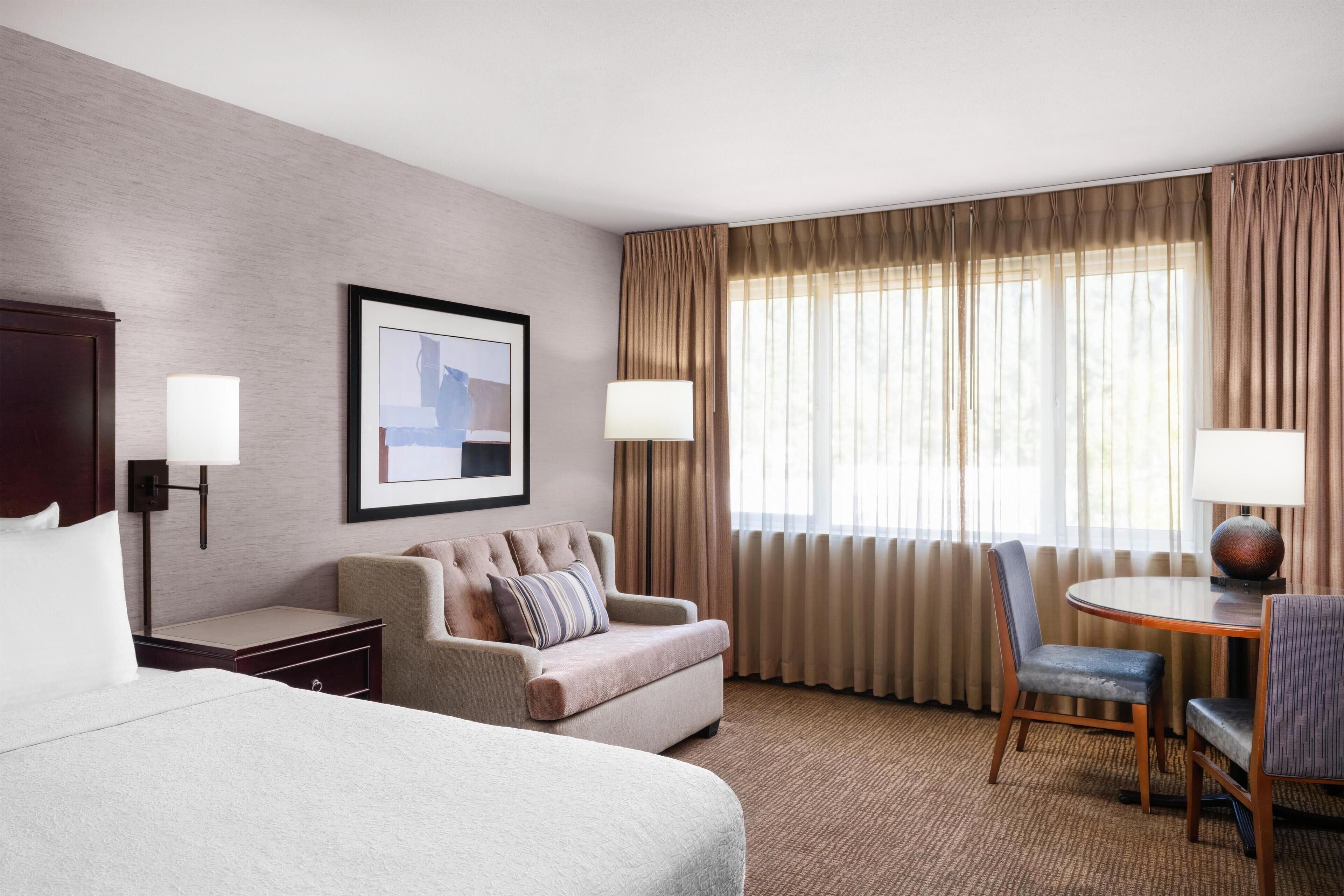 A hotel room with a large bed, a sofa, a dining table with chairs, a lamp, and a framed picture on the wall.