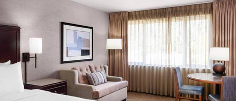 A hotel room with a large bed, a sofa, a dining table with chairs, a lamp, and a framed picture on the wall.
