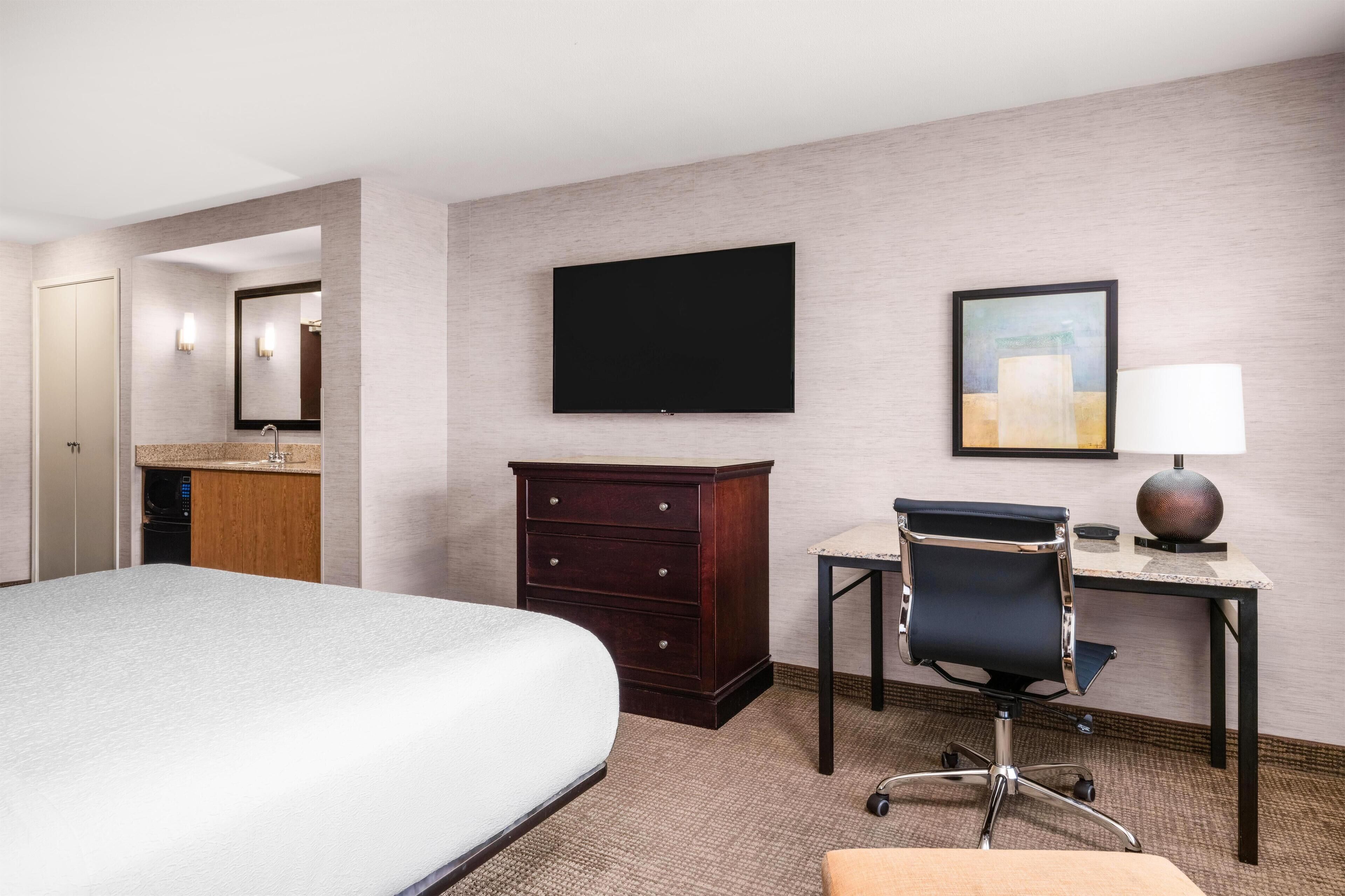 Una habitación de hotel con una cama, un escritorio con silla, un televisor, un aparador y un cuadro en la pared.