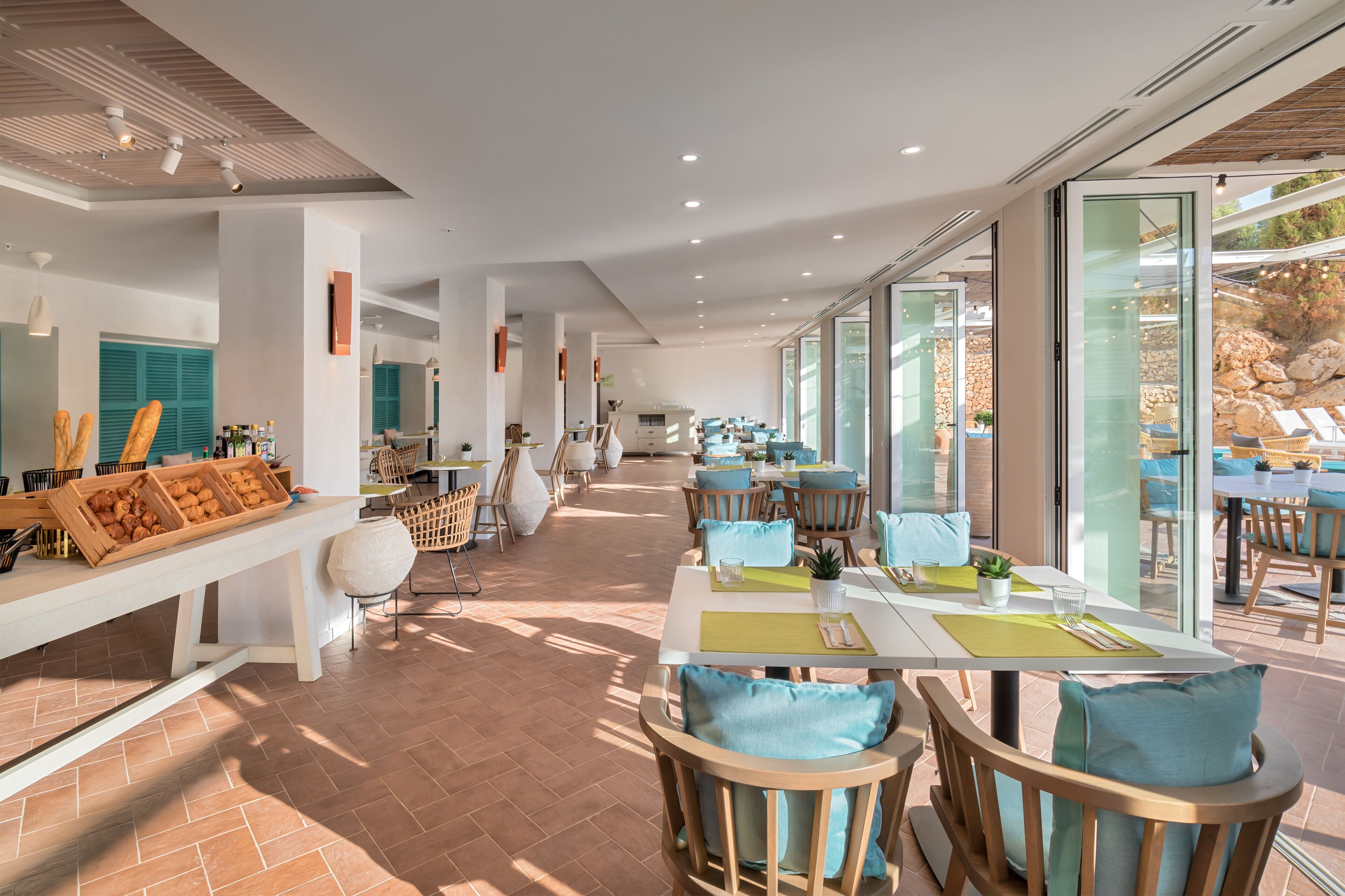 Ein geräumiger Speisebereich mit mehreren Tischen und Stühlen, einem Buffet mit Speisen sowie großen Fenstern mit Blick ins Freie.