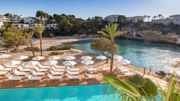 A poolside area with white lounge chairs and umbrellas, a clear blue pool, and a view of the sea and buildings in the background. - Barceló Ponent Beach (Felanitx)