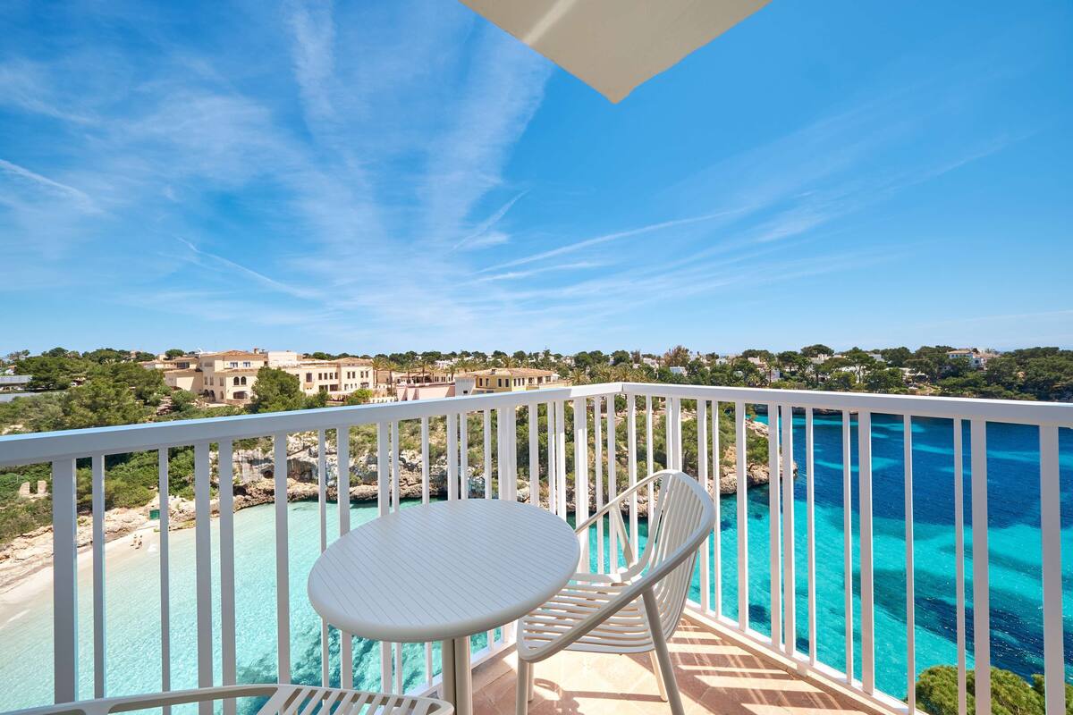 A balcony with a white table and chairs overlooking a clear blue sea and a coastal town.
