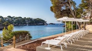 A seaside terrace with white lounge chairs and umbrellas, a rocky coastline, and sailboats in the background. - Barceló Ponent Beach (Felanitx)